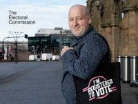 A man is standing outside a train station in a city centre. He has a black tote bag slung over his shoulder. A band-style logo which says “I’m Registered To Vote” can be seen on the bag. 